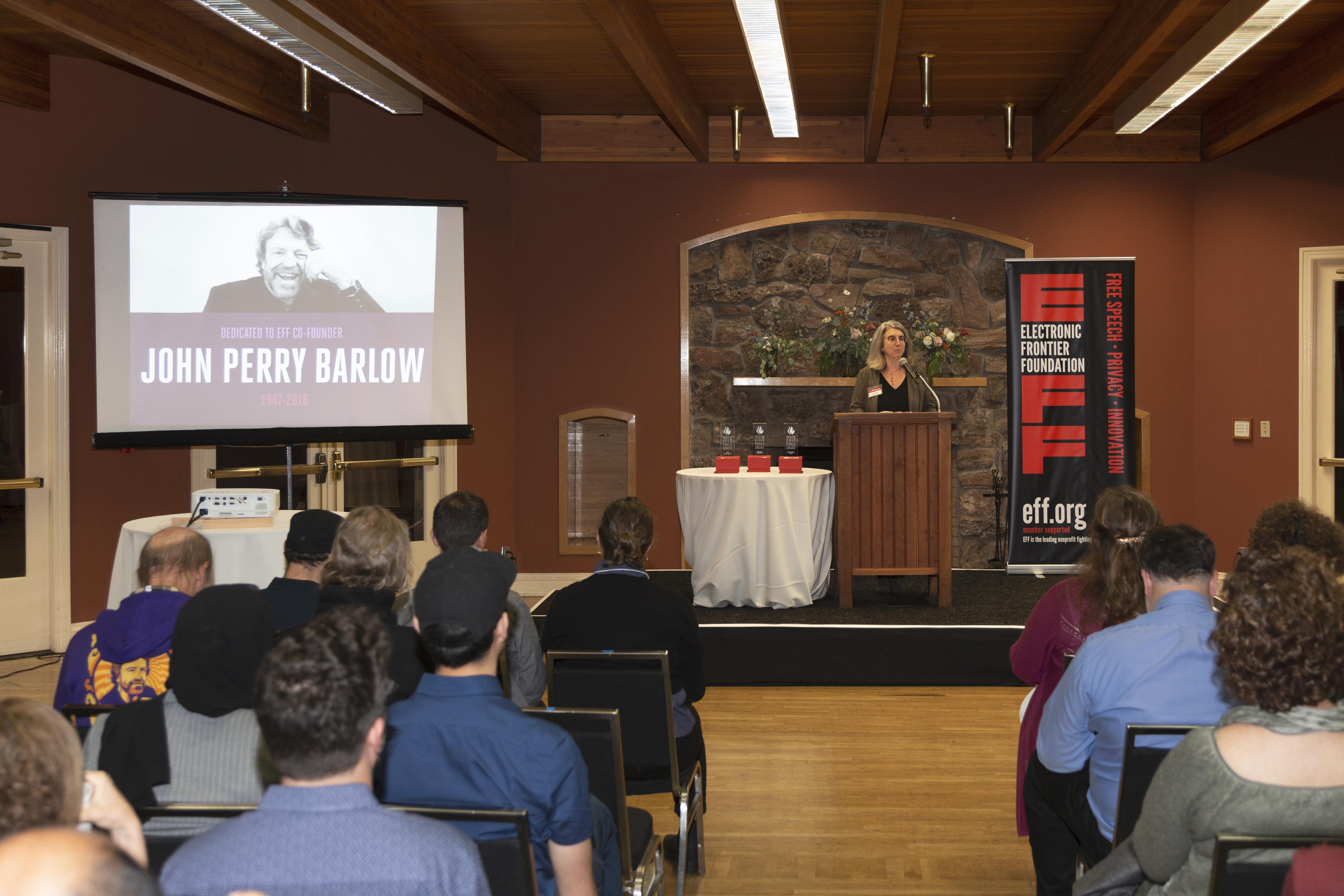 Picture of screen and raised dias with podium. To the left, is a projected image of our co-founder, John Perry Barlow. At the poduim on the right is our Executive Director Cindy Cohn, opening the "Barlow" dedication.