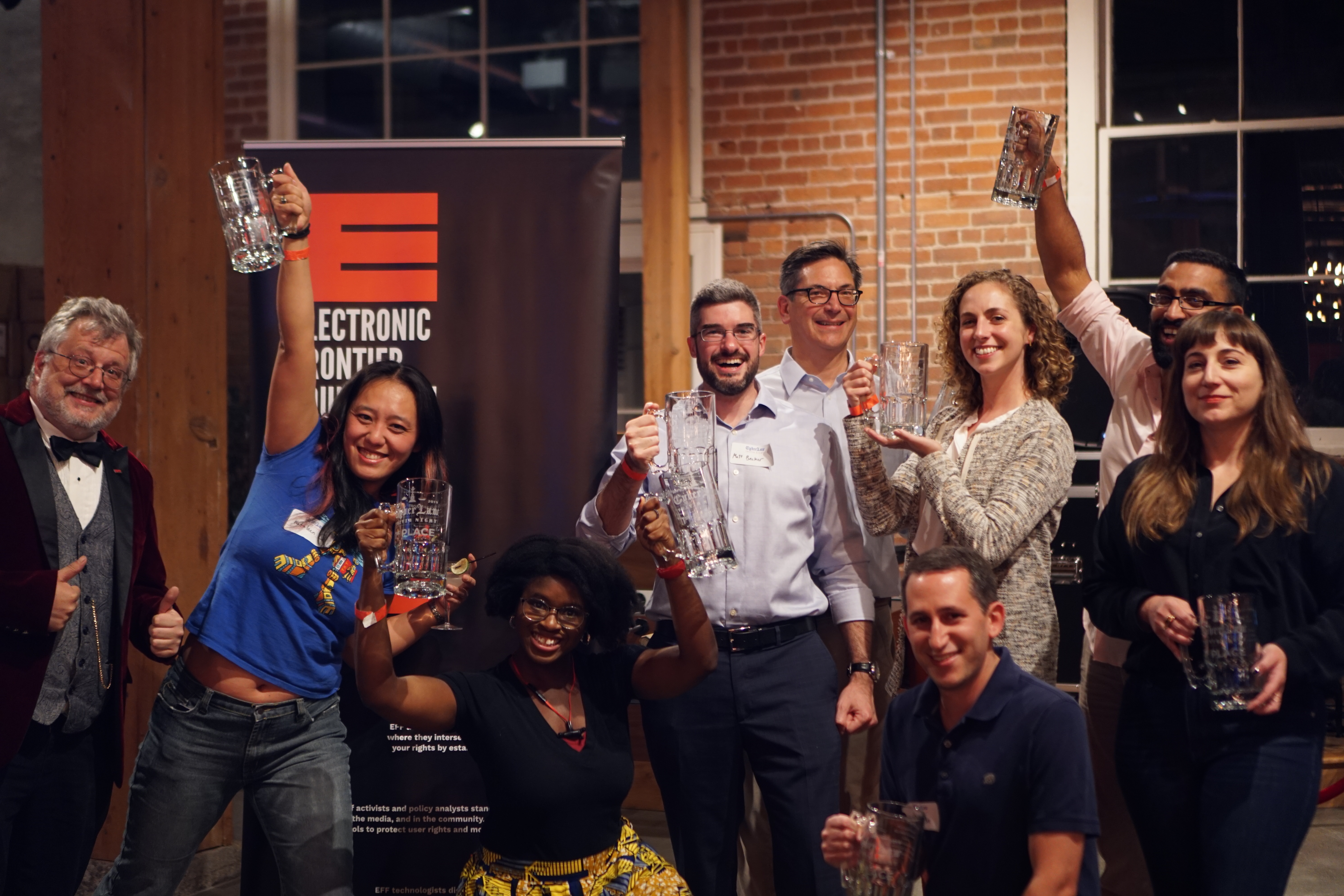  "F.U.C.T. (Fenwick Underwrites Cyberlaw Trivia)" This is a photo of several people holding glass 1st place mugs standing next to an EFF banner, as well as a man in a stylish purple suit—Quizmaster Kurt!