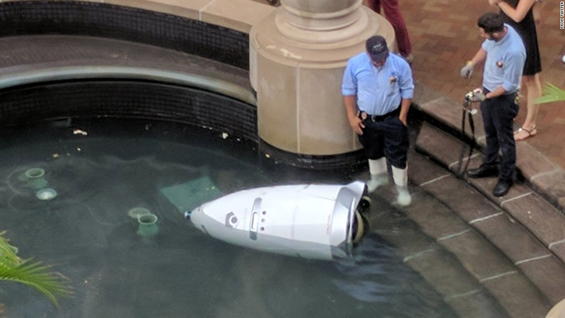 A knightscope surveillance robot lying in a fountain, with two police looking at it quizzically