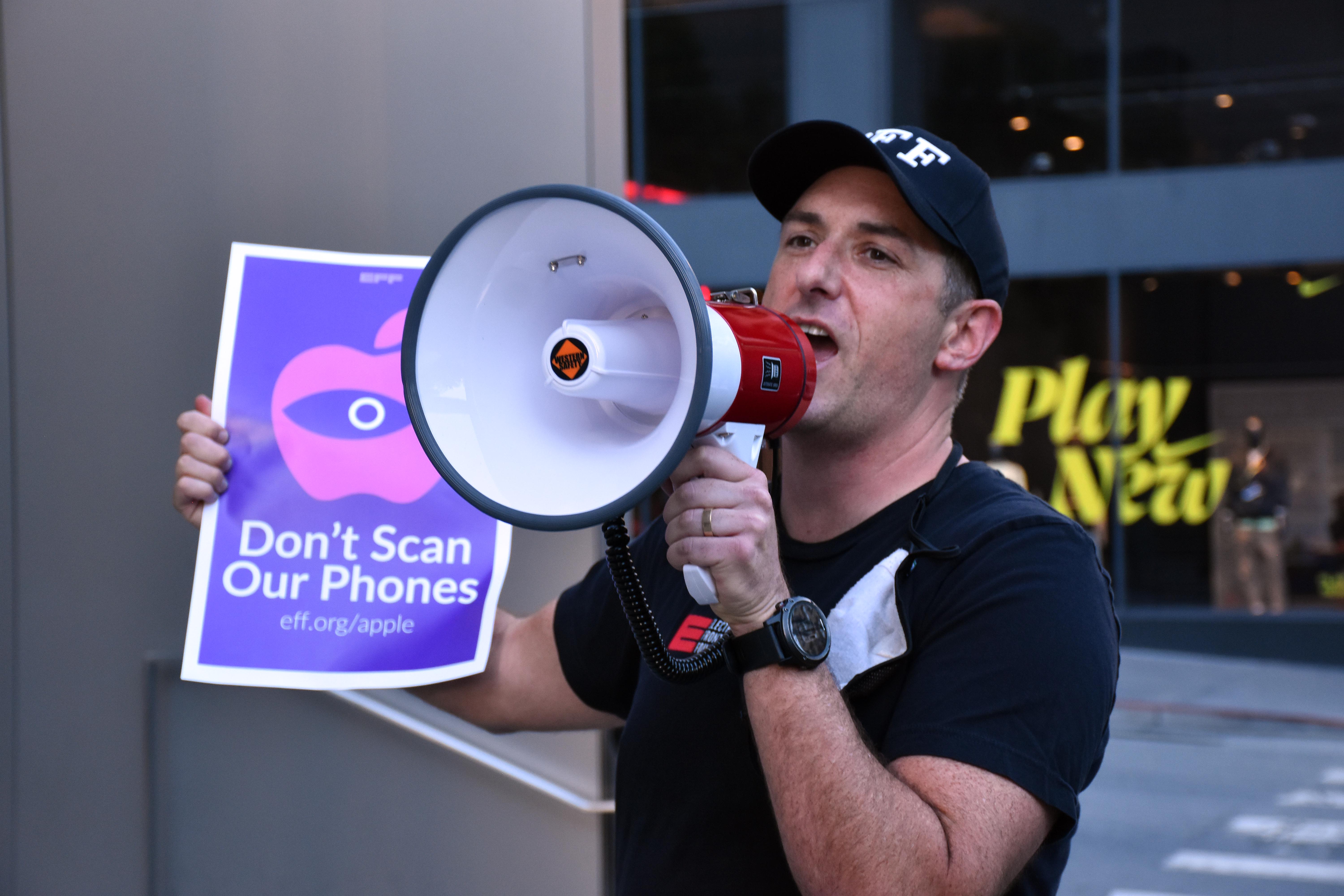 a man wearing an eff hat and shirt holds a bullhorn and a sign reading "don't scan our phones"