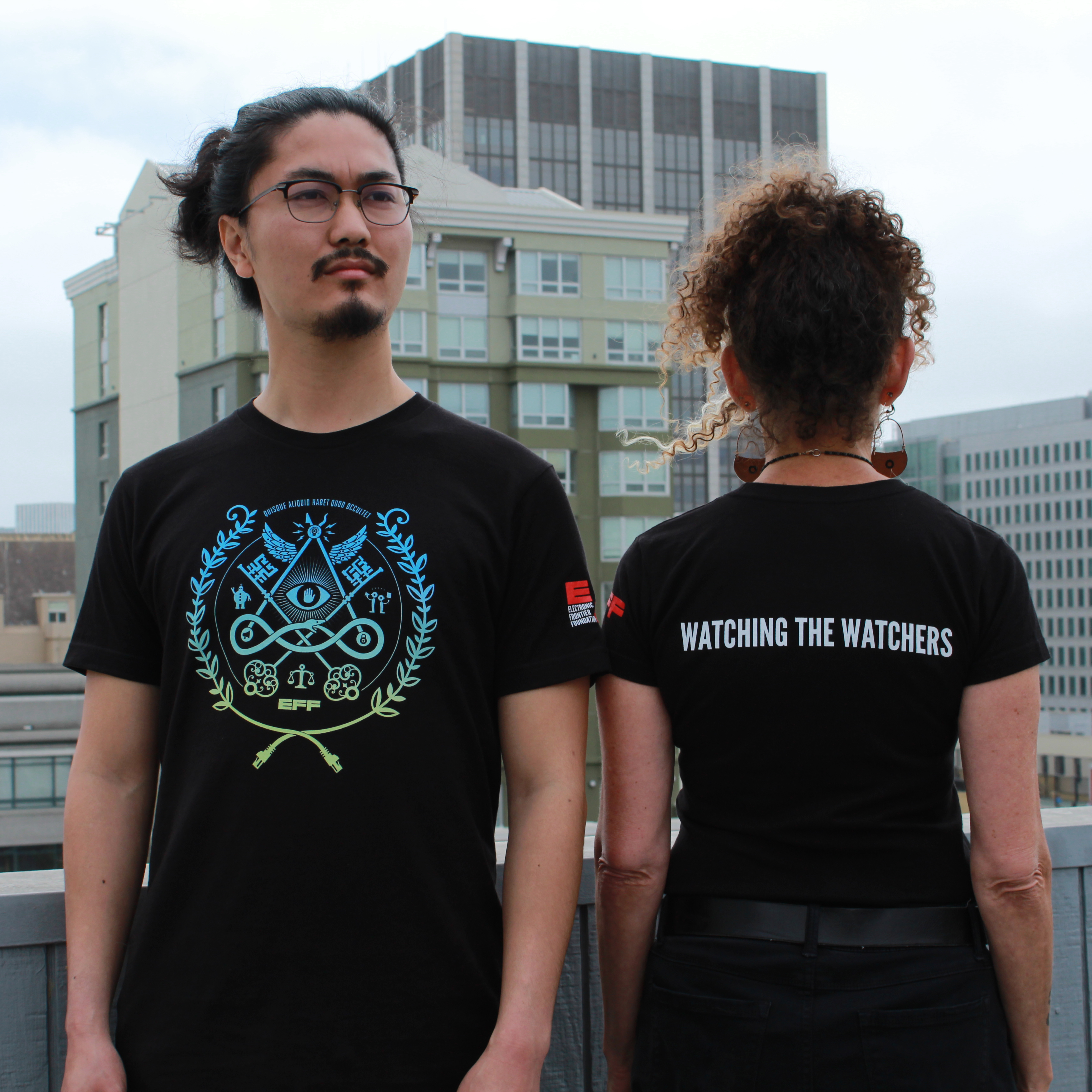 EFF's new member t-shirt. Left person shows the front, which is a privacy focused image with blue to green gradient. Back, shown by right person, says "Watch the Watchers"